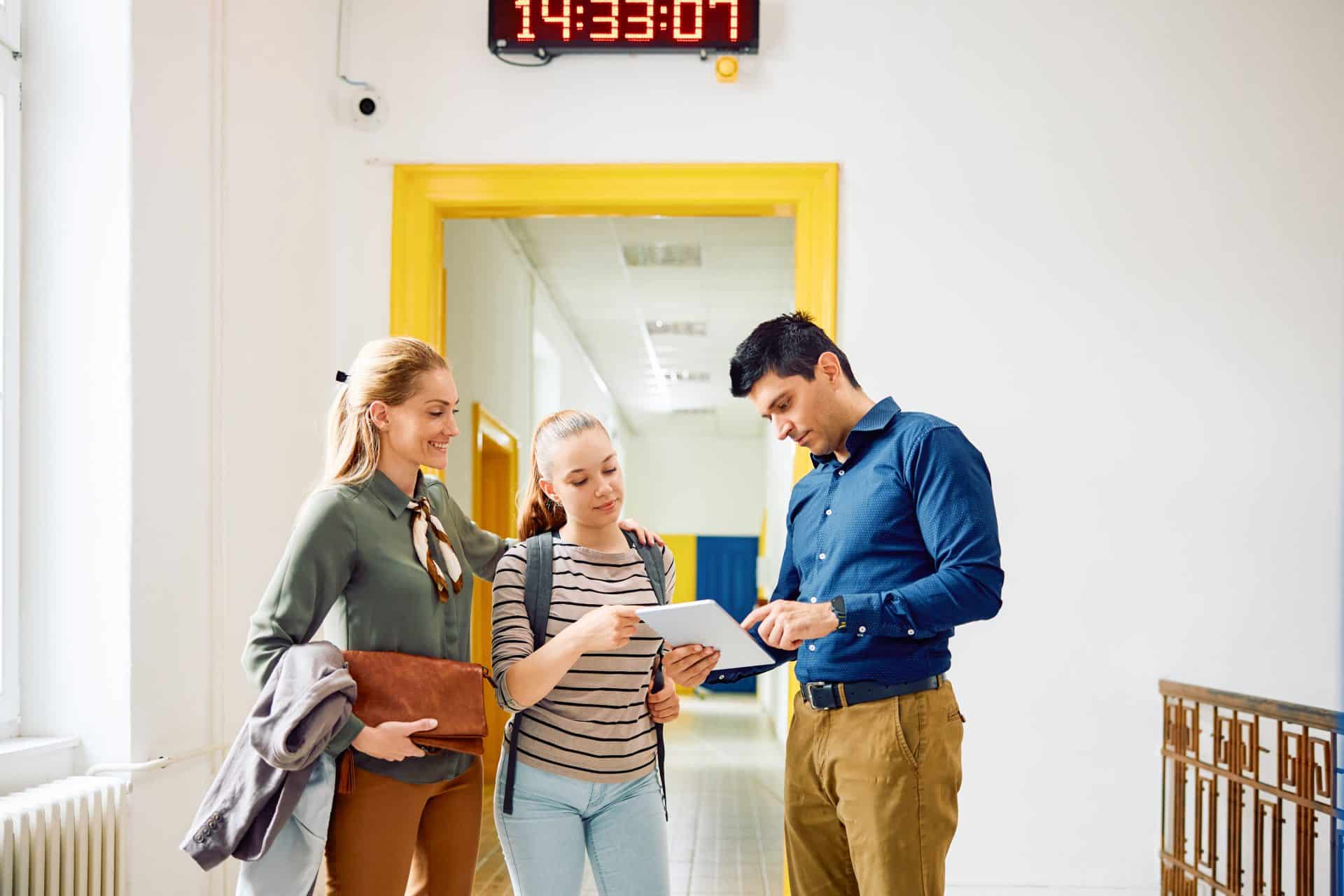 High school teacher using touchpad with his student and her mother in hallway.